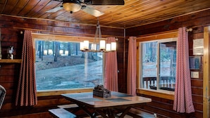 Interior acogedor de una cabaña con una mesa de comedor de madera, bancos, un ventilador de techo y ventanas amplias que ofrecen vistas a un bosque.