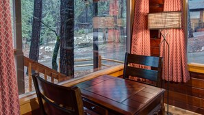Una mesa de madera con sillas, una lámpara y vistas a un bosque a través de amplias ventanas.