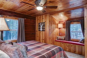 Una habitación tipo cabaña con cama, ventilador de techo, ventana con cortinas y una lámpara de pared.