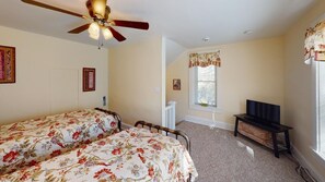 A bedroom with two beds, a ceiling fan, a window with curtains, a television on a stand, and a door leading to another room.