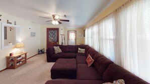 A living room with a sectional sofa, a ceiling fan, and a window with sheer curtains.