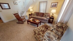 A living room with a brown sofa, a wooden rocking chair, a coffee table, and two lamps. A staircase is visible in the background.