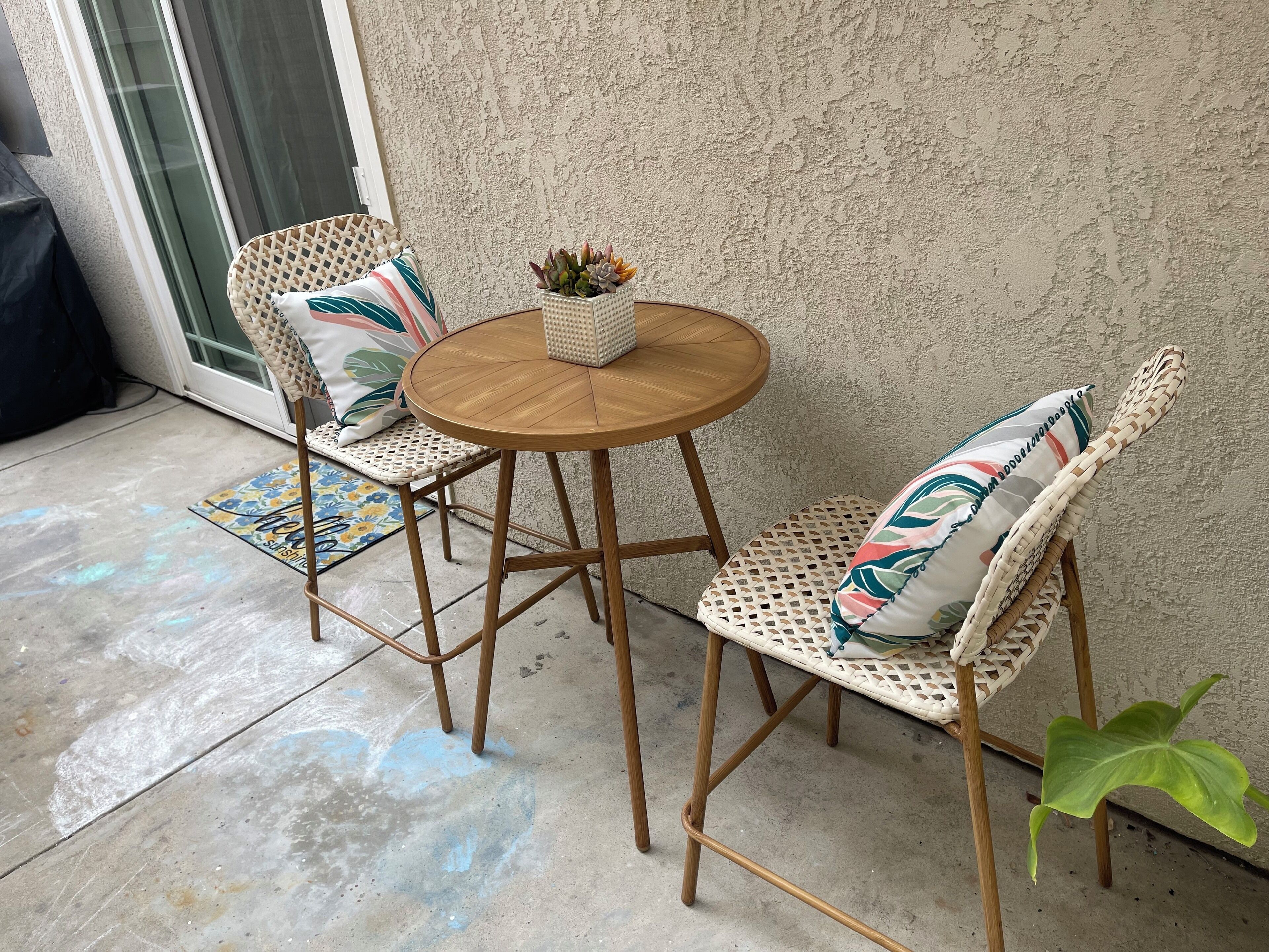 Une table ronde en bois, accompagnée de deux chaises en osier, chacune dotée d’un coussin orné d’un motif de feuilles. Une petite plante en pot est posée sur la table.