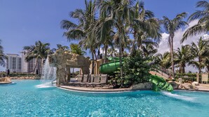 Una piscina con tobogán de agua, palmeras y una estructura rocosa.