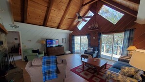 Un salon spacieux avec un plafond haut, des poutres en bois, un ventilateur de plafond, une télévision, un canapé, un fauteuil et un chien.