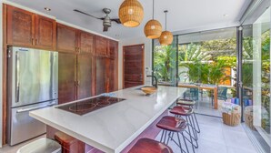 Une cuisine moderne dotée d’un grand îlot central, d’appareils électroménagers en acier inoxydable et d’un coin repas séparé par une paroi de verre, avec un espace extérieur aménagé pour prendre ses repas.