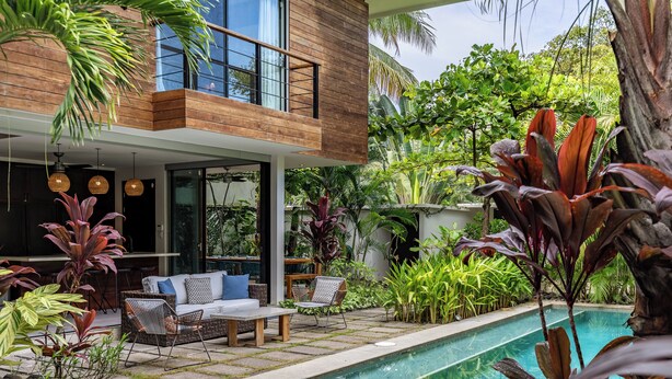 Un espace piscine extérieur moderne, doté d’une terrasse en bois, de chaises longues et d’une végétation tropicale luxuriante.