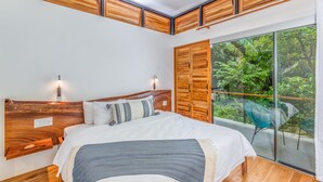Une chambre dotée d’un grand lit, d’une tête de lit en bois et d’une porte-fenêtre coulissante donnant sur un balcon avec vue sur la verdure.
