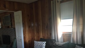 Una sala de estar acogedora con un sofá gris oscuro, cojines estampados y piso de madera. Se ve una puerta blanca y una ventana con cortinas.