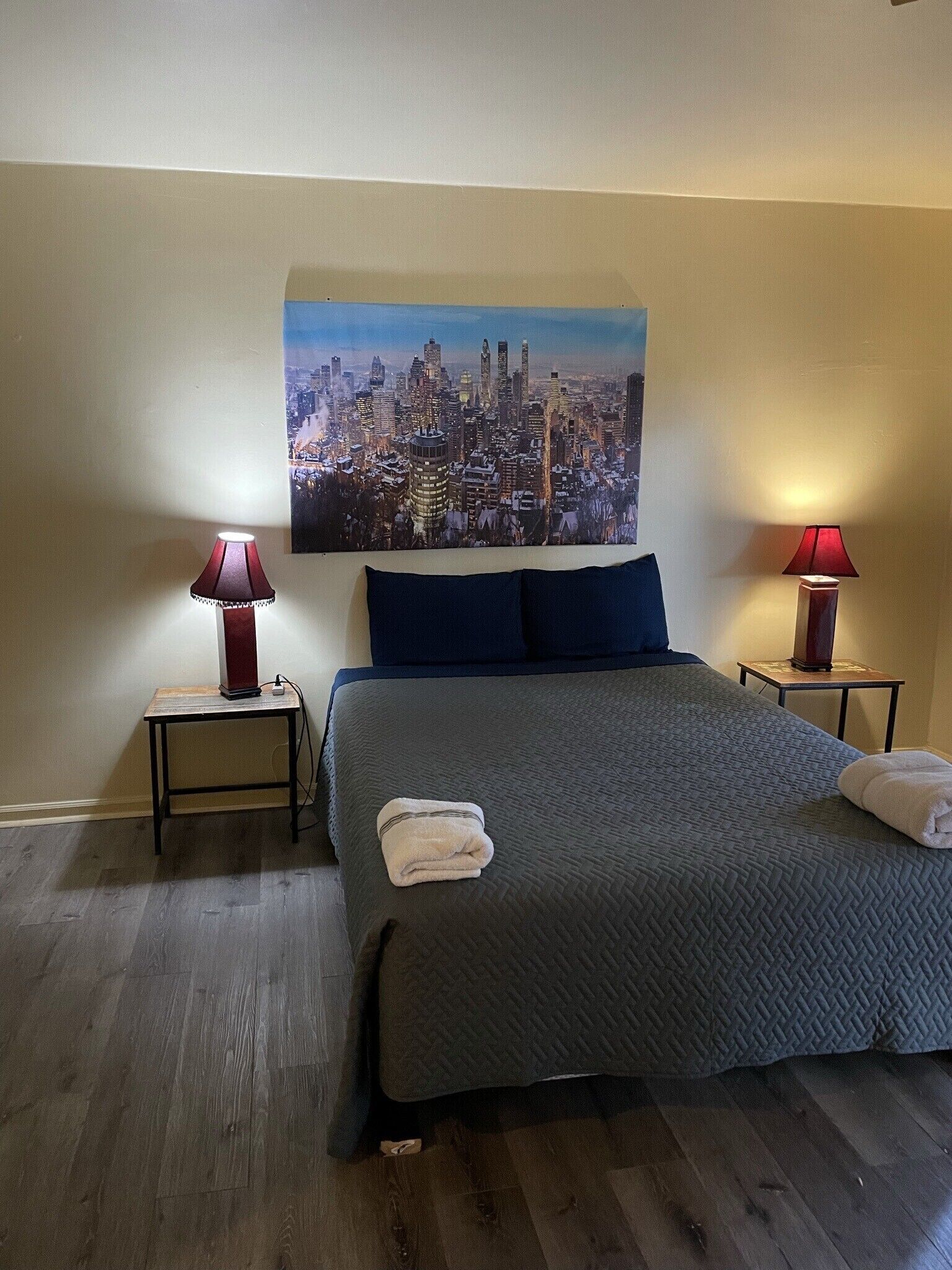 Ein Hotelzimmer mit einem großen Bett, zwei Nachttischen, zwei Lampen, einem Stadtbild an der Wand und einem ordentlich bezogenen Bett mit Handtüchern.