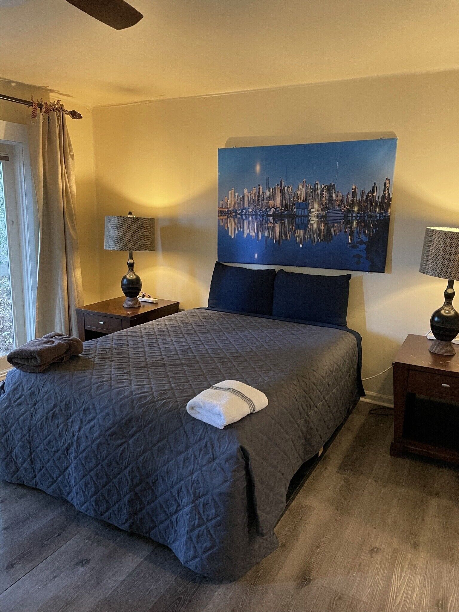 A hotel room with a large bed, two bedside tables with lamps, a cityscape painting on the wall, and a window with curtains.