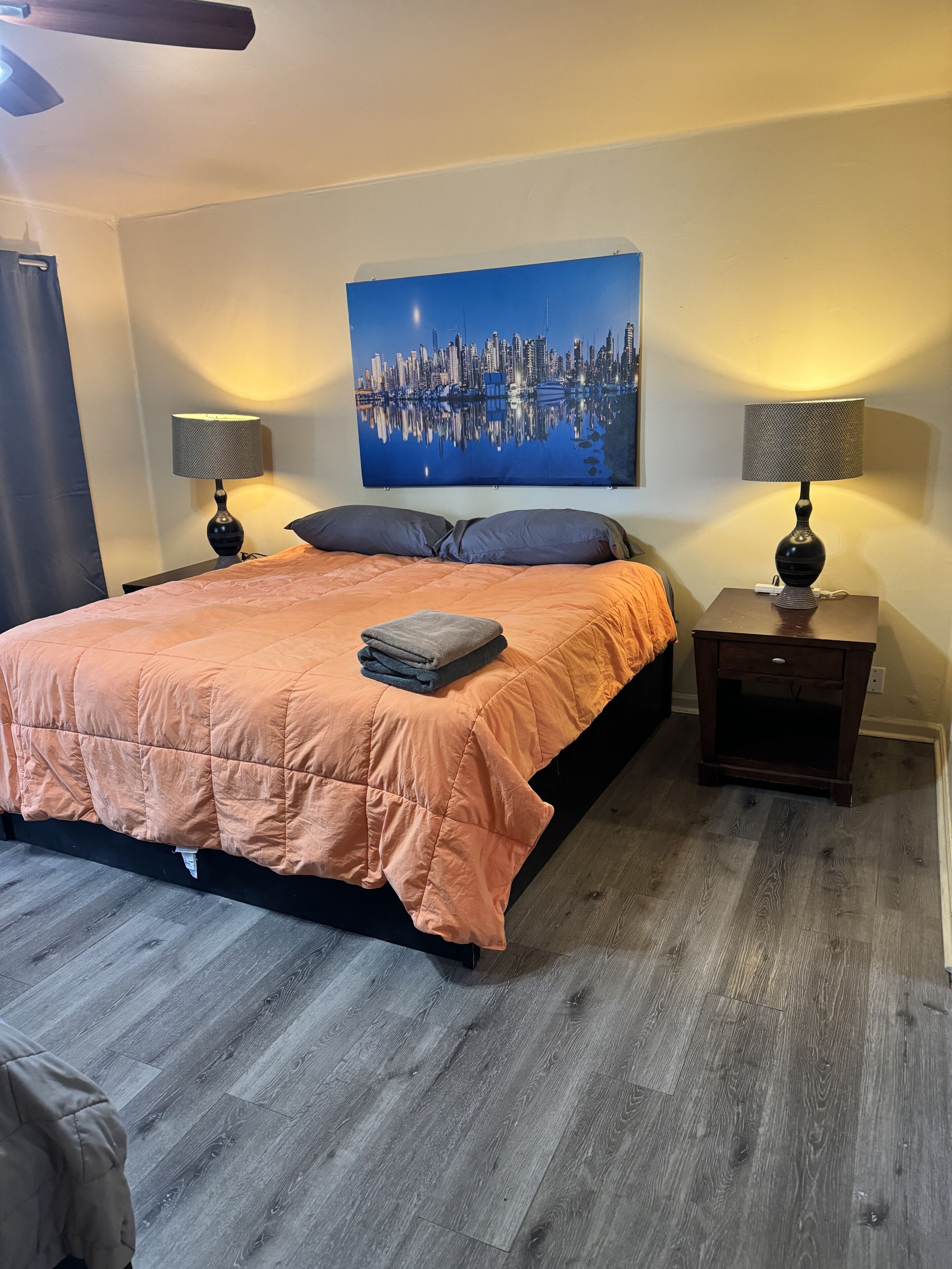 Een slaapkamer met een groot bed, twee nachtkastjes met lampen, een stadslandschap op de muur, en een houten vloer.
