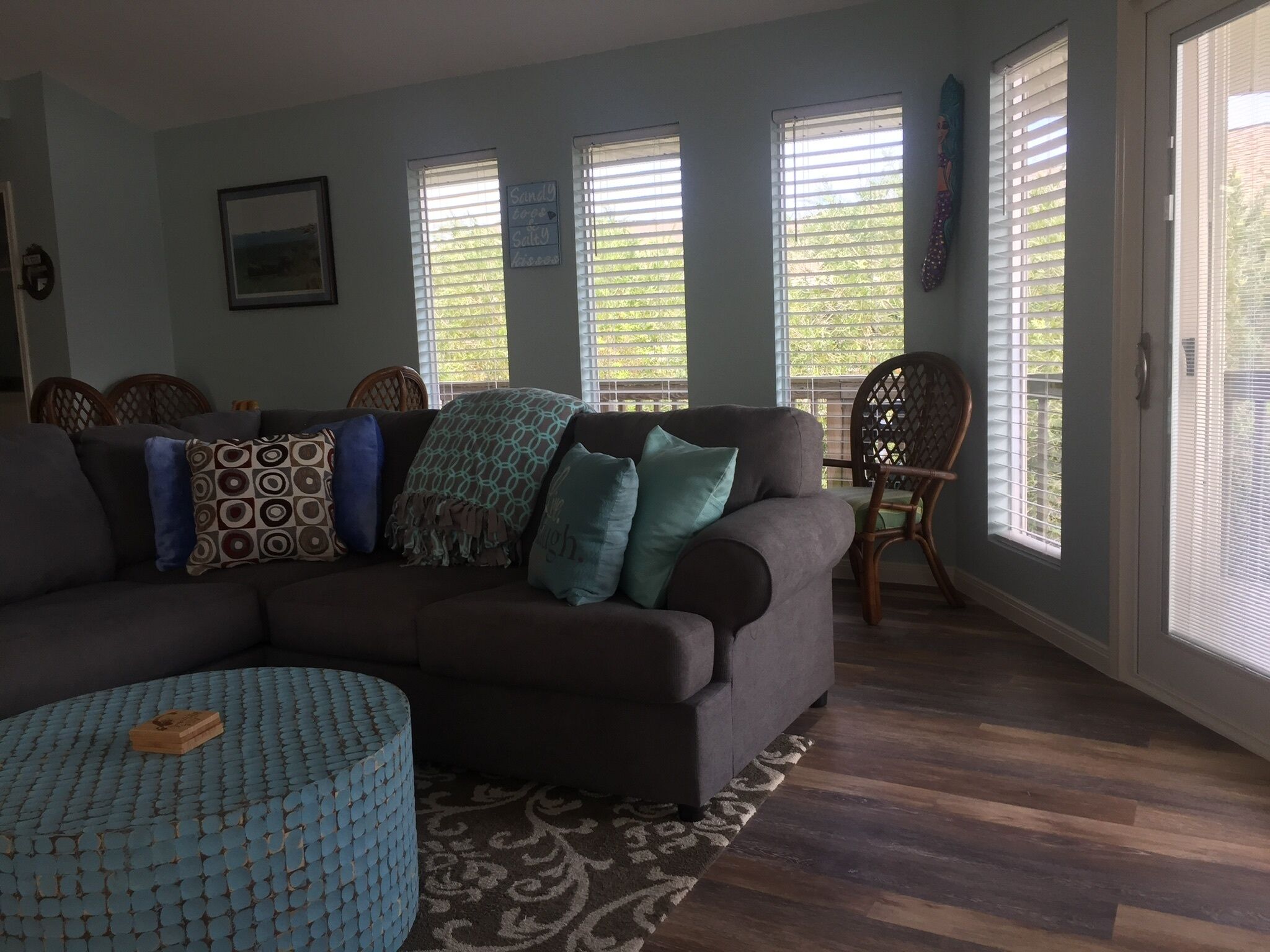 A living room with a sectional sofa, patterned cushions, a blue ottoman, and a patterned rug. There are two windows with blinds, a chair, and a wall-mounted sign.