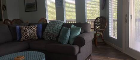 A living room with a sectional sofa, patterned cushions, a blue ottoman, and a patterned rug. There are two windows with blinds, a chair, and a wall-mounted sign.