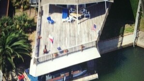 Vista aérea de um barco atracado com deck coberto, cadeiras de descanso e uma bandeira.