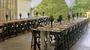 Une longue table à manger, dressée pour un repas, entourée de chaises, avec des rideaux blancs et des compositions florales en guise de décoration centrale.