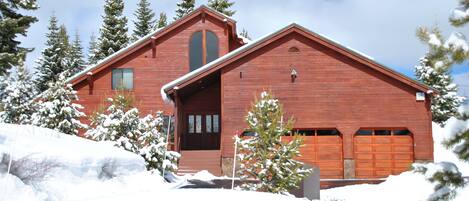 A red wooden cabin with a large garage, surrounded by snow-covered trees and a clear sky.
