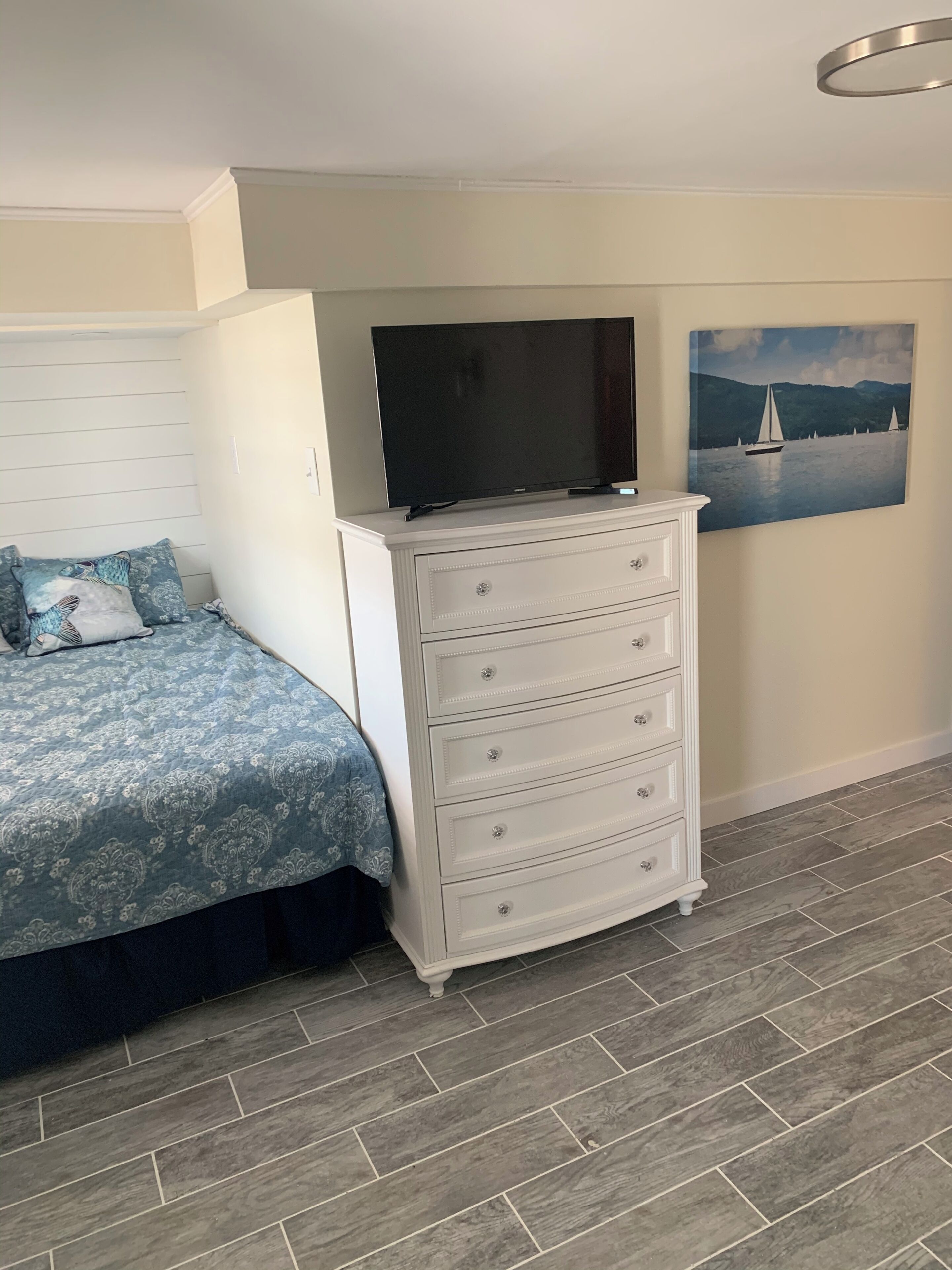Une chambre à coucher avec un lit, une commode, un téléviseur et un tableau accroché au mur.