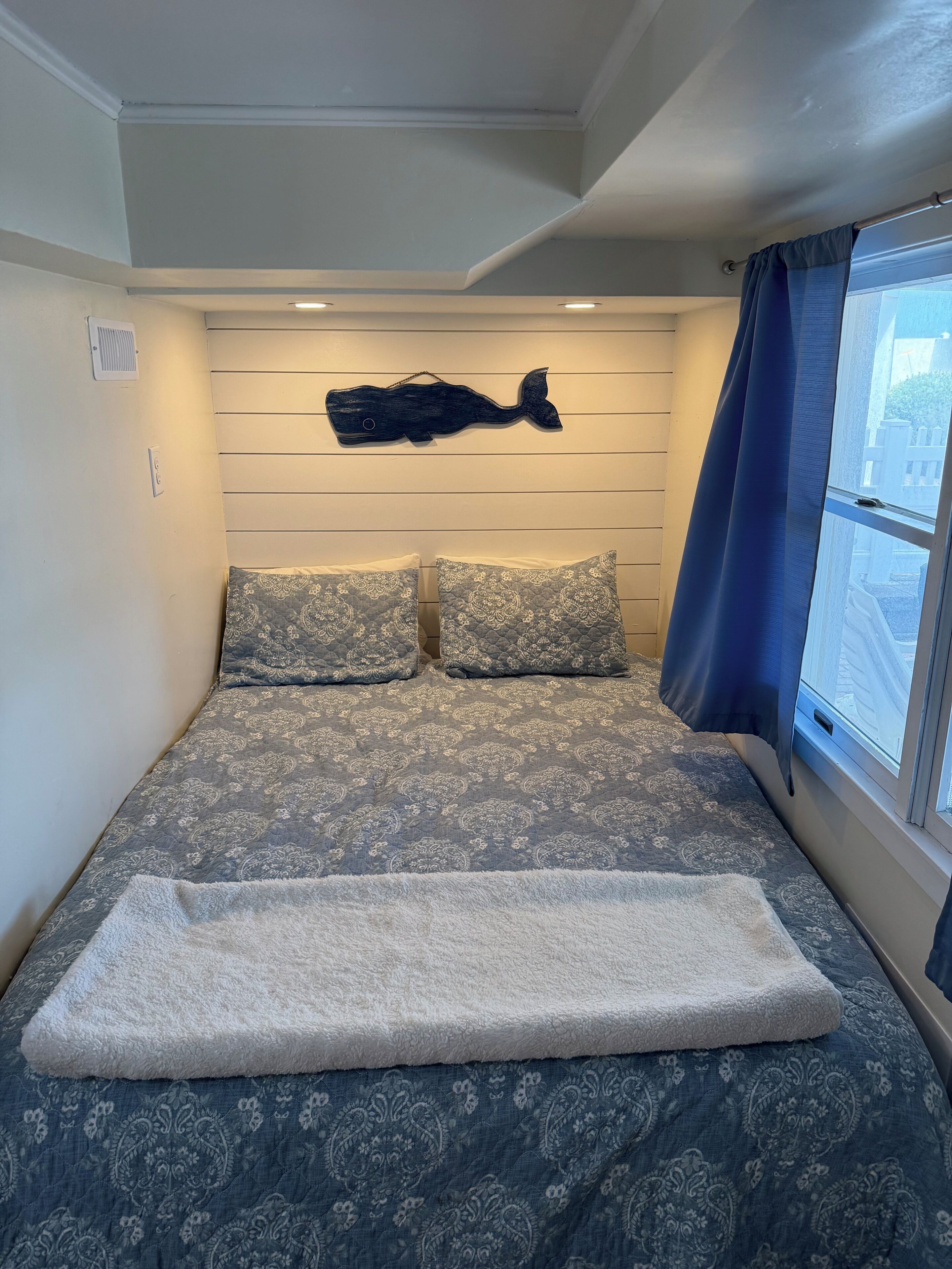A bedroom with a bed, two patterned pillows, a dark wall decoration, a window with a curtain, and a white headboard.