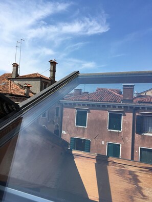 A view from a window overlooking a street with terracotta-tiled buildings, a clear blue sky, and a visible antenna on a building.