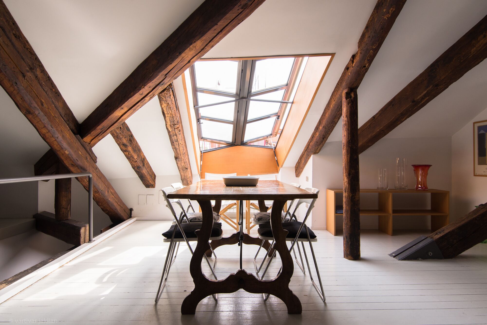Une pièce mansardée aux plafonds hauts, avec des poutres apparentes, une lucarne et une table centrale entourée de chaises.