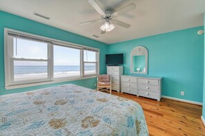Un dormitorio con una cama grande, un aparador, una silla, un ventilador de techo, un televisor y vista al océano a través de una ventana.