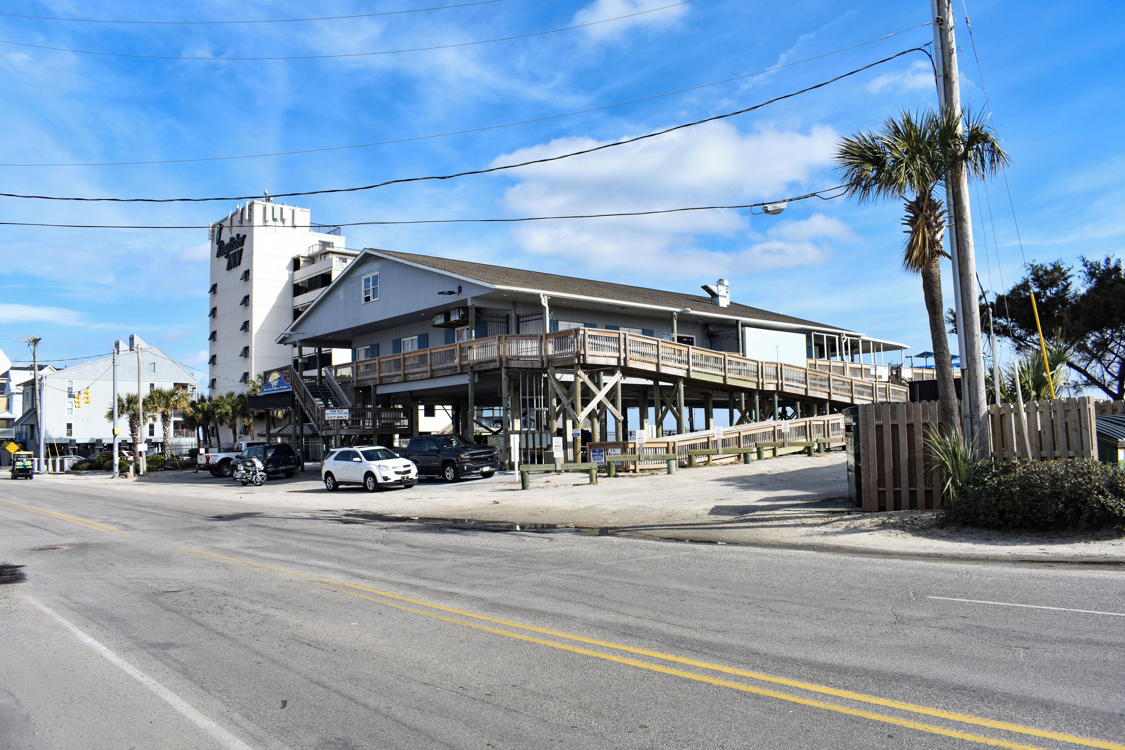 A coastal building with a wooden deck, a staircase leading to the upper floor, and a parking area with cars.