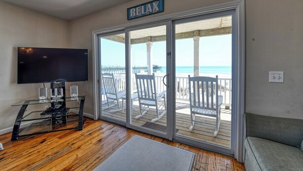 A living room with a TV, speakers, and a glass door leading to a balcony with chairs and a 'RELAX' sign. - Against The Wind A, Oceanfront Home, Amazing Pier View, Duplex (Garden City Beach)