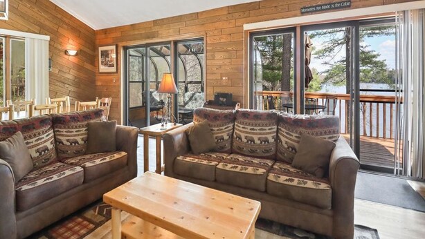 A cozy living room with a wooden interior, a large sofa, a coffee table, and a sliding glass door leading to a deck. - Executive Year Round Home ~ Minutes From  Minocqua (Arbor Vitae)