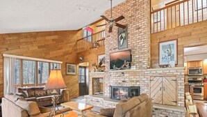 A spacious living area with a fireplace, wooden walls, and a loft bedroom with a balcony. - Executive Year Round Home ~ Minutes From  Minocqua (Arbor Vitae)