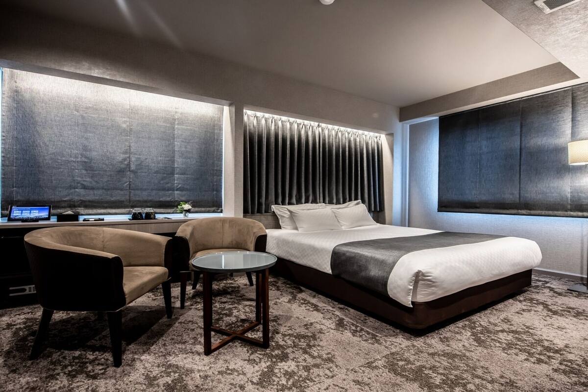 Una habitación de hotel con una cama, un escritorio con computadora, una silla, una mesita y una lámpara.