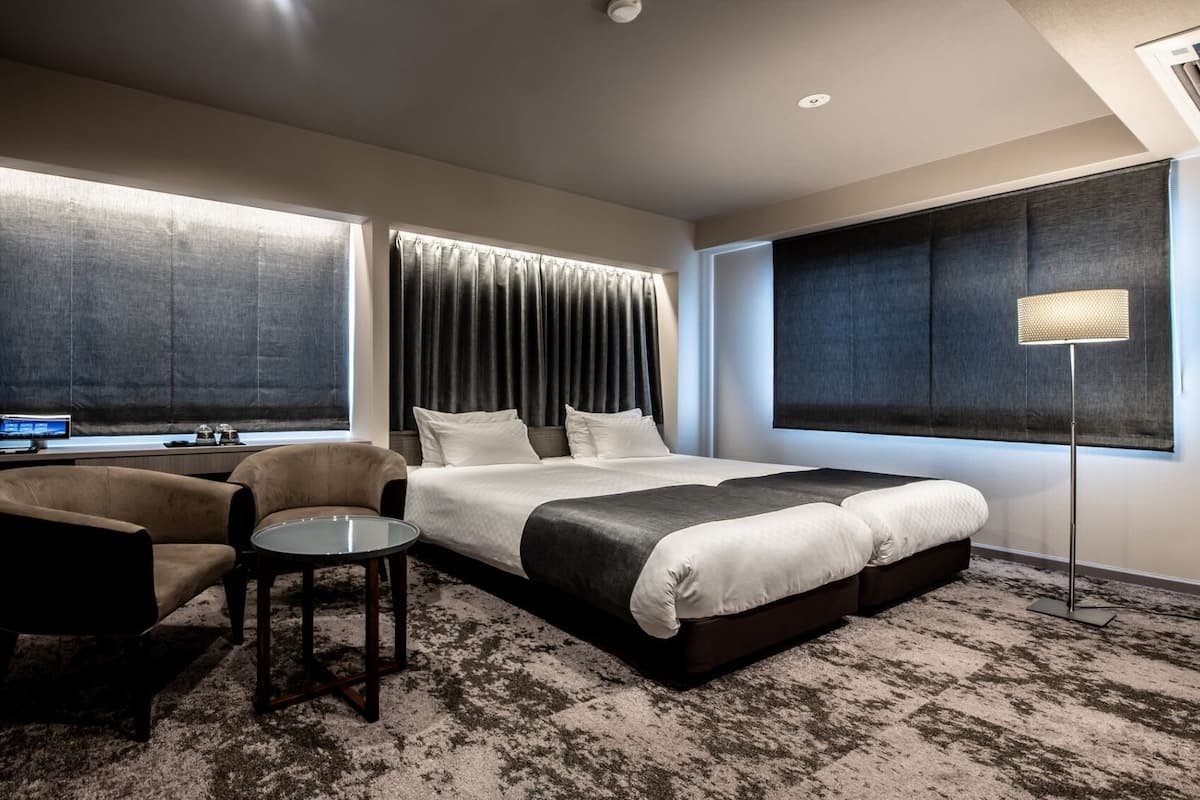 Quarto de hotel com cama, secretária com computador, cadeira, mesinha pequena e abajur.