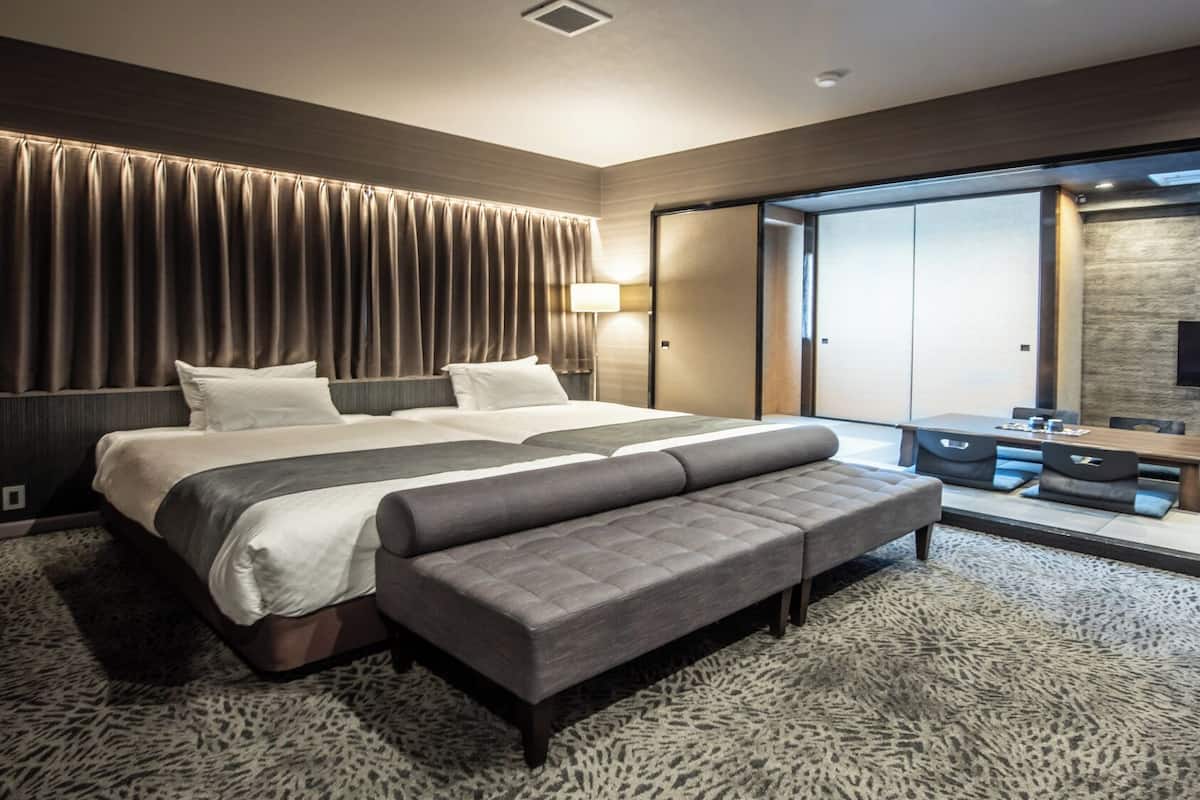 Quarto de hotel moderno com uma cama grande, um banco, uma TV e uma porta deslizante para uma varanda.