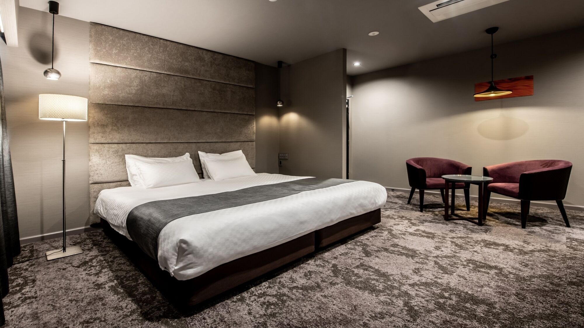 A modern hotel room with a large bed, a seating area with two chairs, and a floor lamp.
