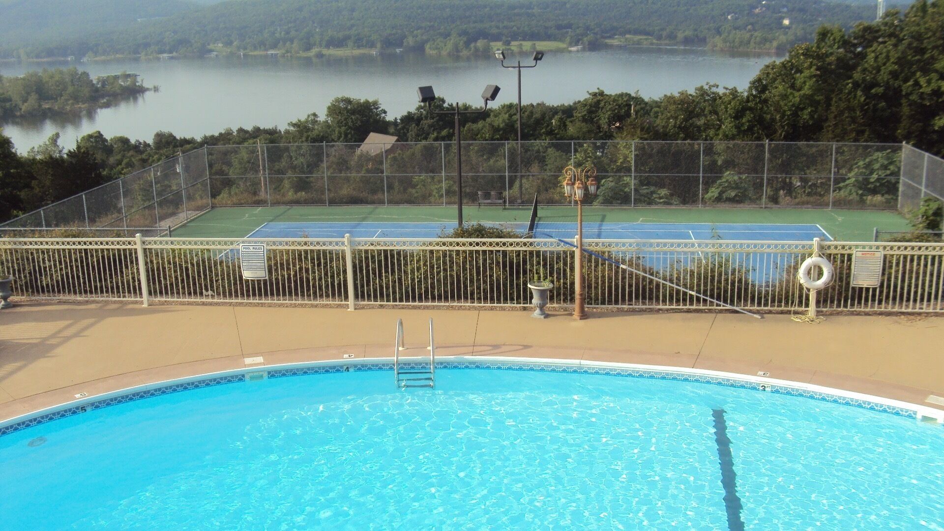 A swimming pool with a view of a tennis court and a lake in the background.