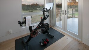 Een stilstaande fiets met een digitaal scherm, een handdoek en een paar schoenen op een mat, met uitzicht op een landschap door grote ramen.