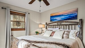 A bedroom with a rustic wooden headboard, a large window with curtains, a painting of a sunset, and a bedside lamp. - Bella Vista~ 5 Star ~ Clean Cabin ~Hot Tub and Beautiful View ~Pets Allowed (Maggie Valley)