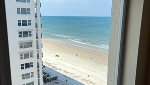 Vista desde una ventana sobre una playa con piscina y un rascacielos.