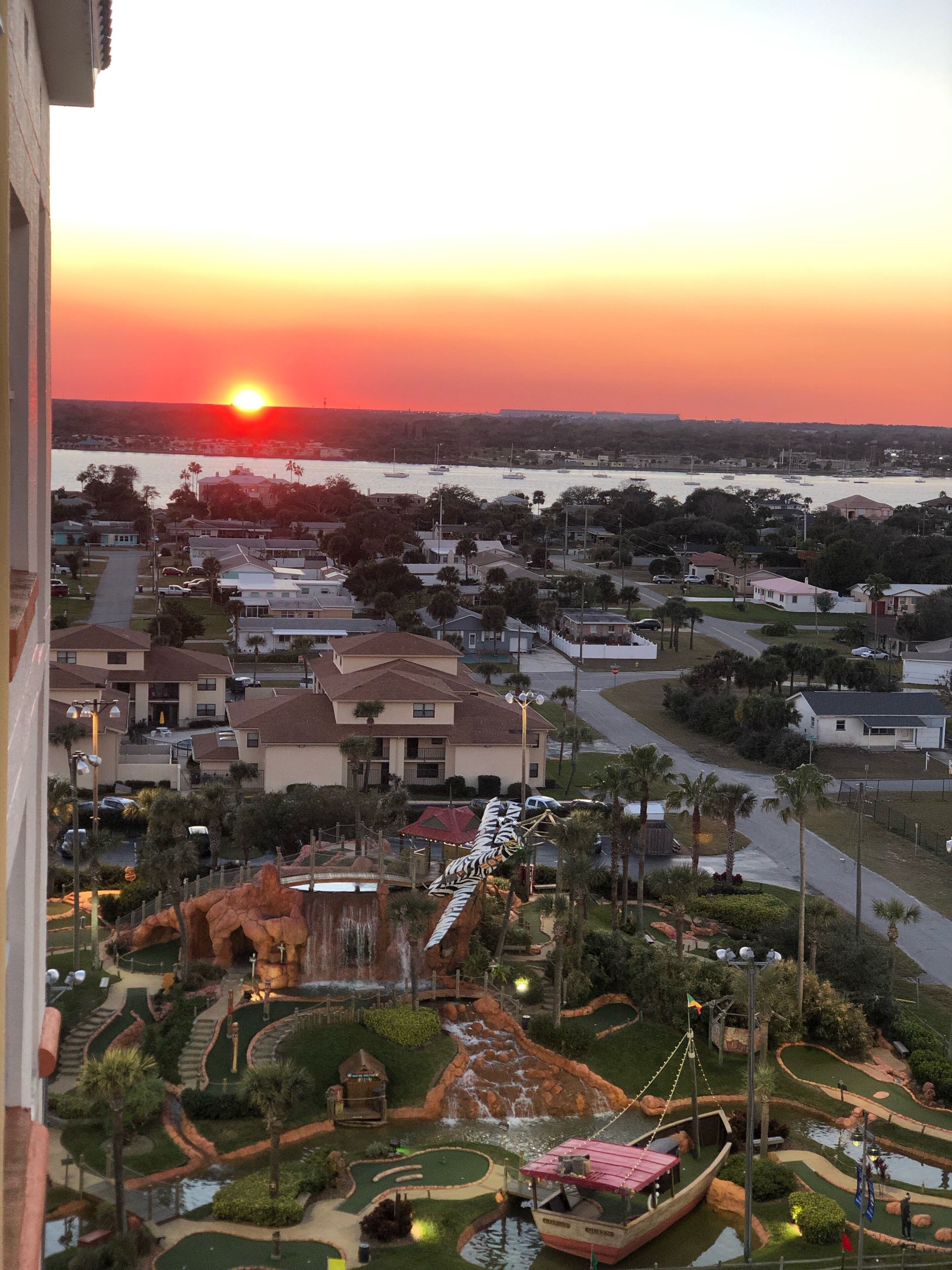 Un quartier résidentiel avec des maisons, un petit parc agrémenté d’un point d’eau et un coucher de soleil sur une étendue d’eau.