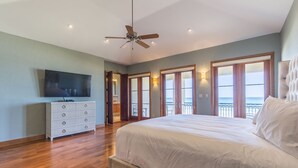 Une chambre spacieuse avec un grand lit, un plancher en bois, un ventilateur de plafond, une télévision et un balcon offrant une vue sur l’océan.