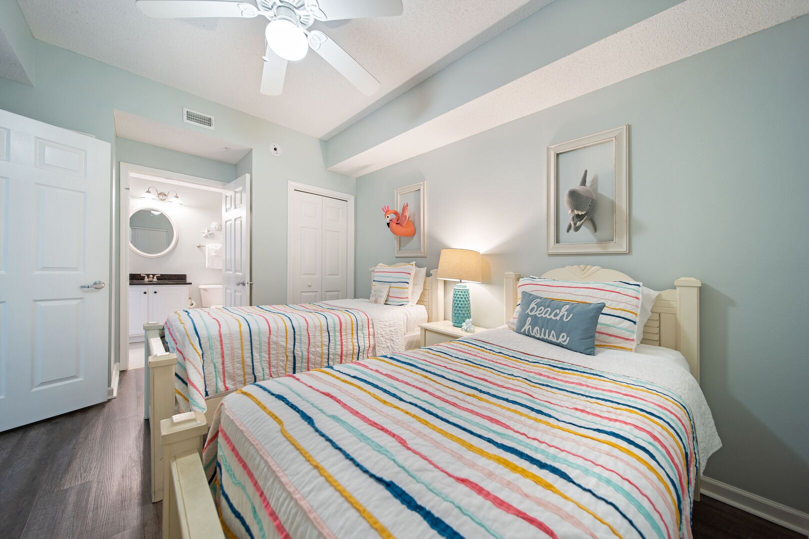 A bedroom with two beds, a ceiling fan, a bathroom with a round mirror, and a beach-themed pillow.