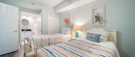 A bedroom with two beds, a ceiling fan, a bathroom with a round mirror, and a beach-themed pillow.
