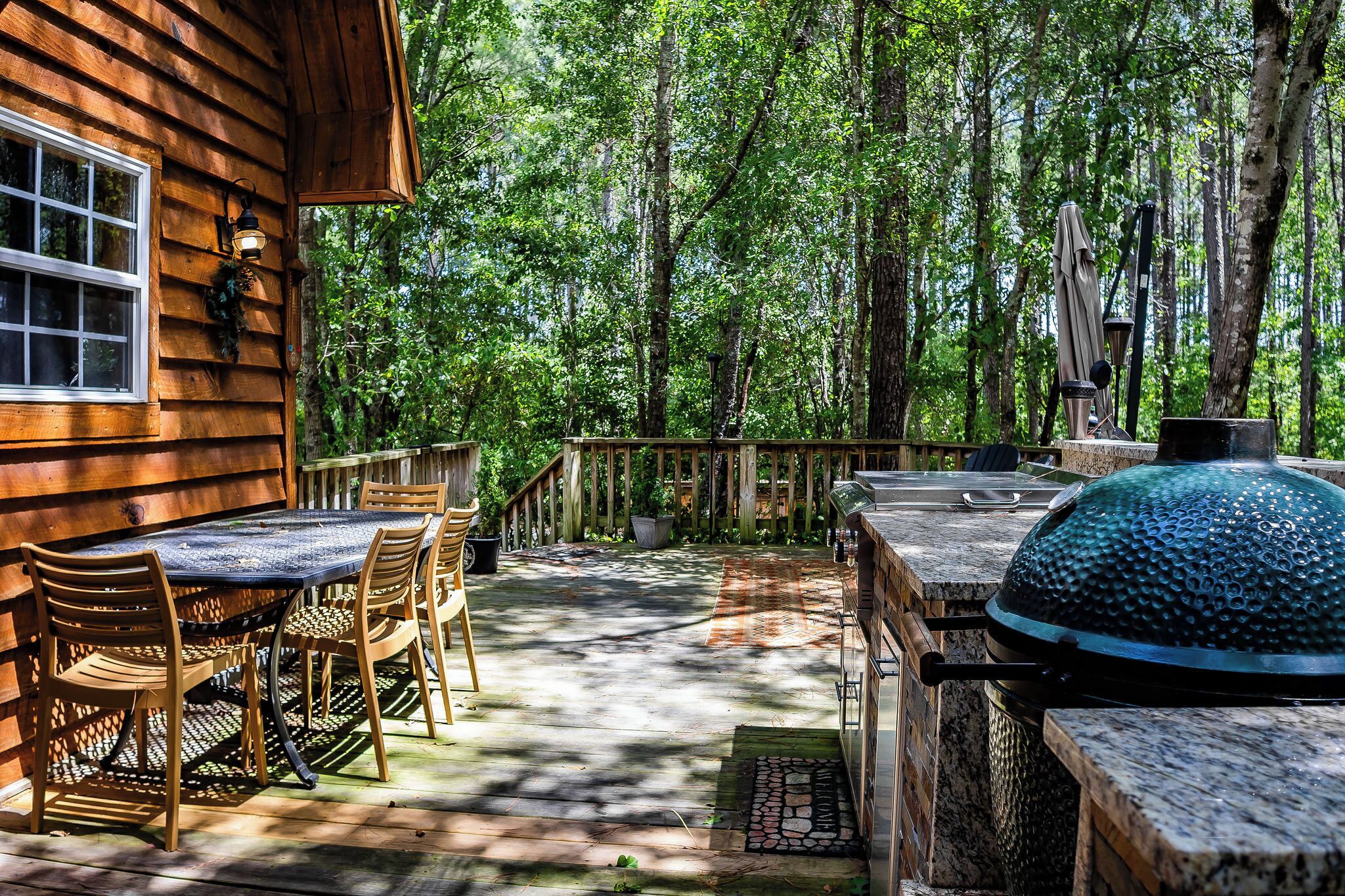 A wooden deck with a table and chairs, a grill, and a window overlooking a forest.