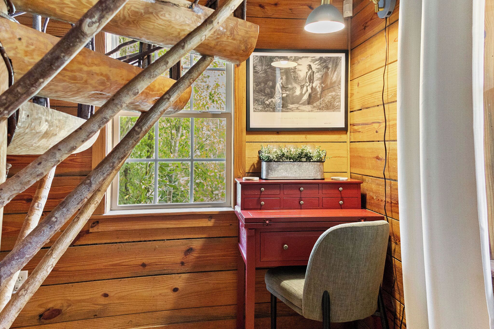 A wooden interior with a staircase, a desk with drawers, a window with a view, and a framed picture on the wall.