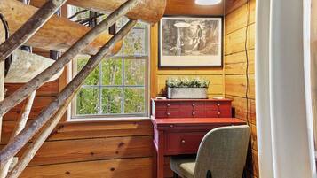 A wooden interior with a staircase, a desk with drawers, a window with a view, and a framed picture on the wall.