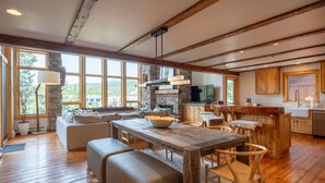 Un vaste espace de vie comprenant une table à manger en bois, des sièges confortables, une cheminée en pierre et de grandes fenêtres offrant une vue sur l’extérieur.