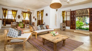 Amplia sala de estar con un sofá de madera, una mesa de centro y una mesa de comedor. Hay cortinas con un estampado tropical, una lámpara de techo tejida y vistas a la vegetación exterior.