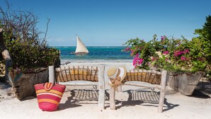 Un paisaje de playa con un velero, sillas de mimbre, un bolso a rayas y plantas en macetas.