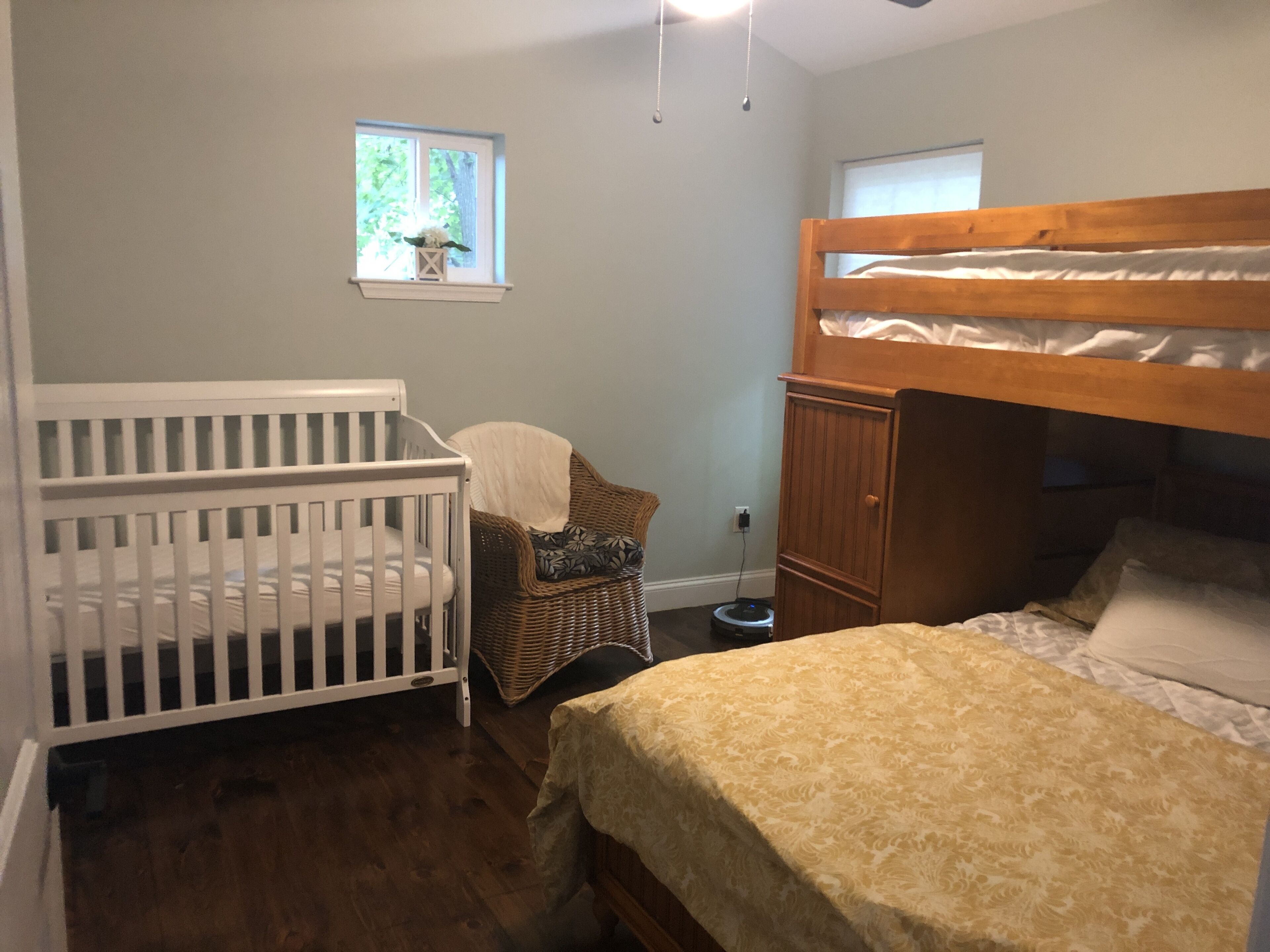 A room with a crib, a small table with a chair, a bunk bed, and a window with a view of greenery.