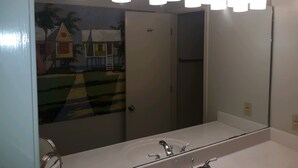 A bathroom with a large mirror, a sink, and a shower. There is a painting on the wall depicting a house and palm trees.